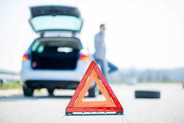 24 x 7 Assistance A young man with a silver car that broke down on the road.He has set up a warning triangle.He is waiting for the technician to arrive.
